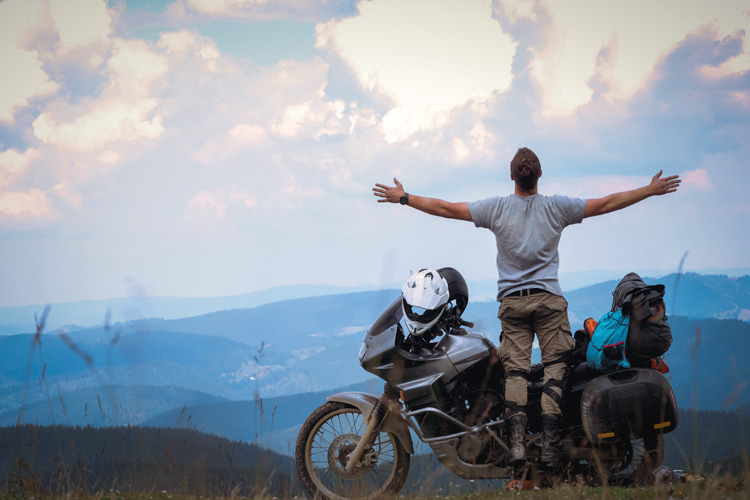 Un hombre de pie al lado de una motocicleta con las dos manos en cruz mientras mira el paisaje de una montaña.