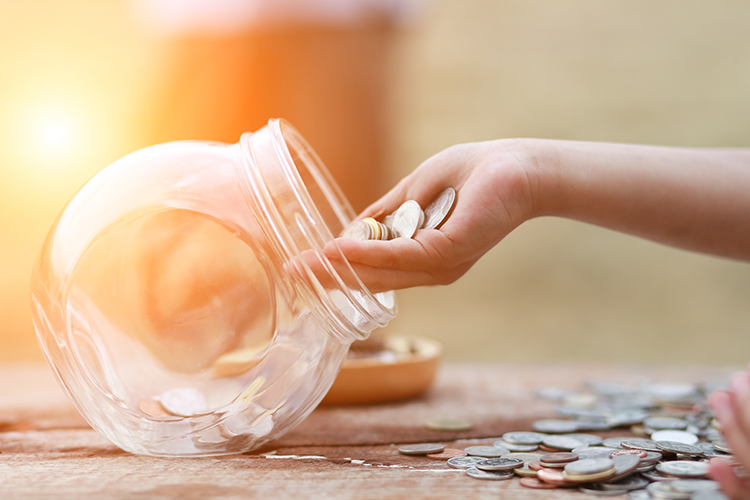 Una mano introduciendo monedas en un bote de cristal, de fondo hay una mesa de madera y el sol.