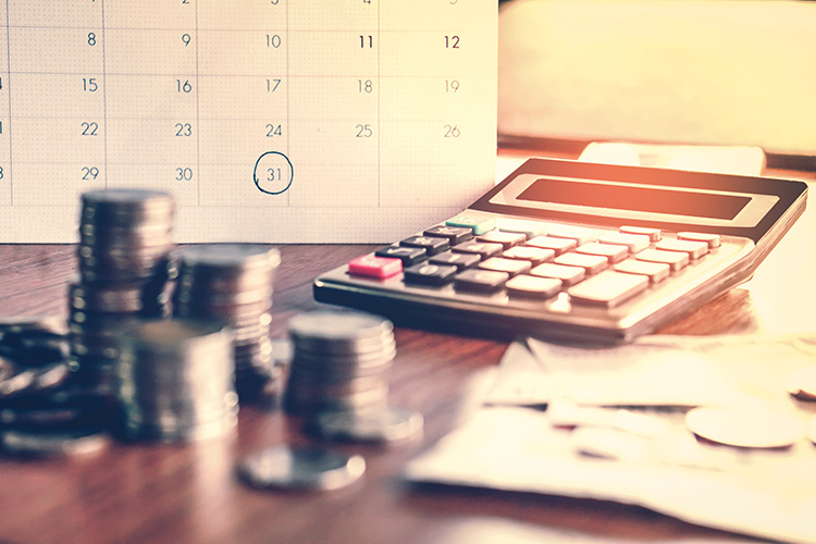 Una calculadora, varias pilas de monedas y un calendario con el número 31 marcado encima de una mesa de madera.