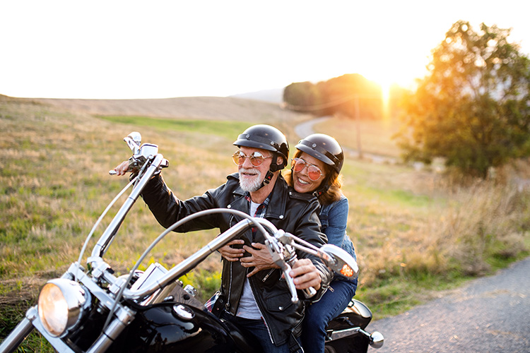 Una pareja con cascos negros iguales viaja en moto de gran cilindrada por una carretera de campo con el fondo de sol.