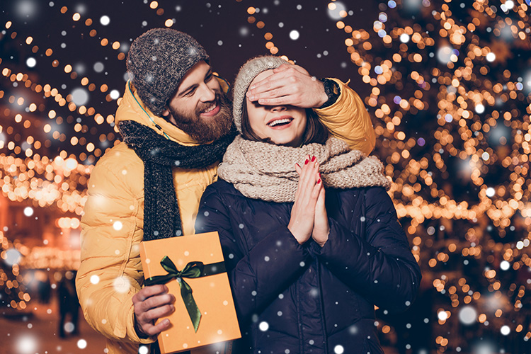 Un hombre con un abrigo amarillo le da un regalo a una mujer con gorro mientras le tapa los ojos con la otra mano.