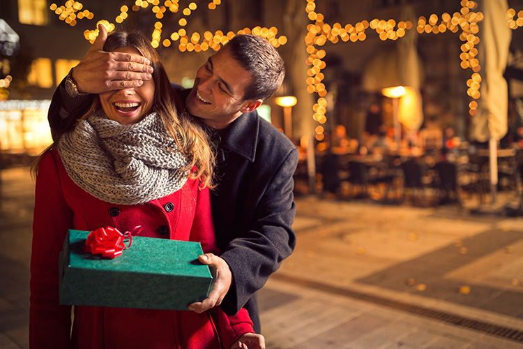 Un hombre le da un regalo a una mujer mientras que le tapa los ojos con la mano derecha, de fondo una terraza borrosa.