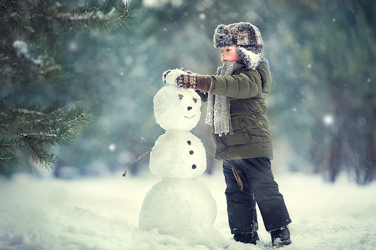 Un niño con guantes y un gorro con orejeras hace un muñeco de nieve con unos árboles verdes desenfocados de fondo