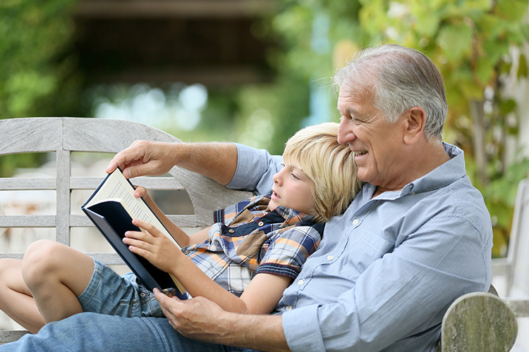 Un abuelo con camisa azul leyendo a su nieto un libro en un banco de madera en un jardín con hojas verdes