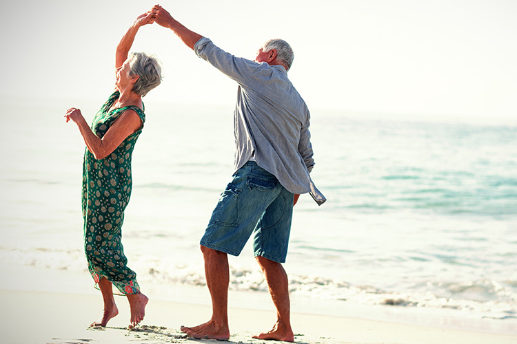 Un hombre y una mujer de avanzada edad bailan por la playa descalzos en la orilla de la playa en un día soleado.