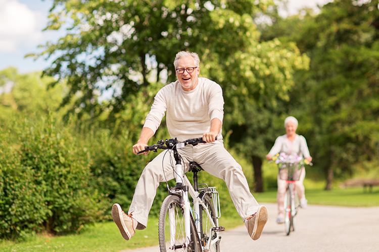 Un hombre de mediana edad monta sonriente en su bicicleta por un jardín, de fondo, una mujer monta en su bicicleta.